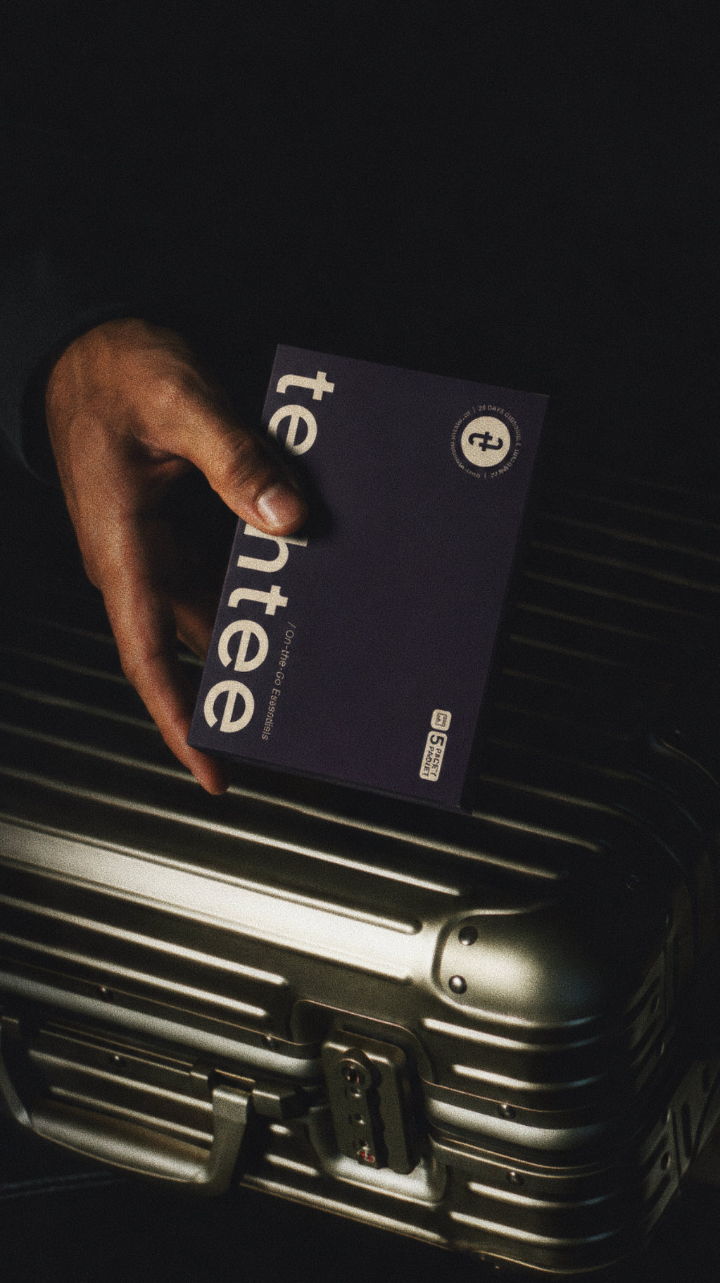 Hand holding a box of 'techtee' travel underwear over a silver suitcase on a dark background