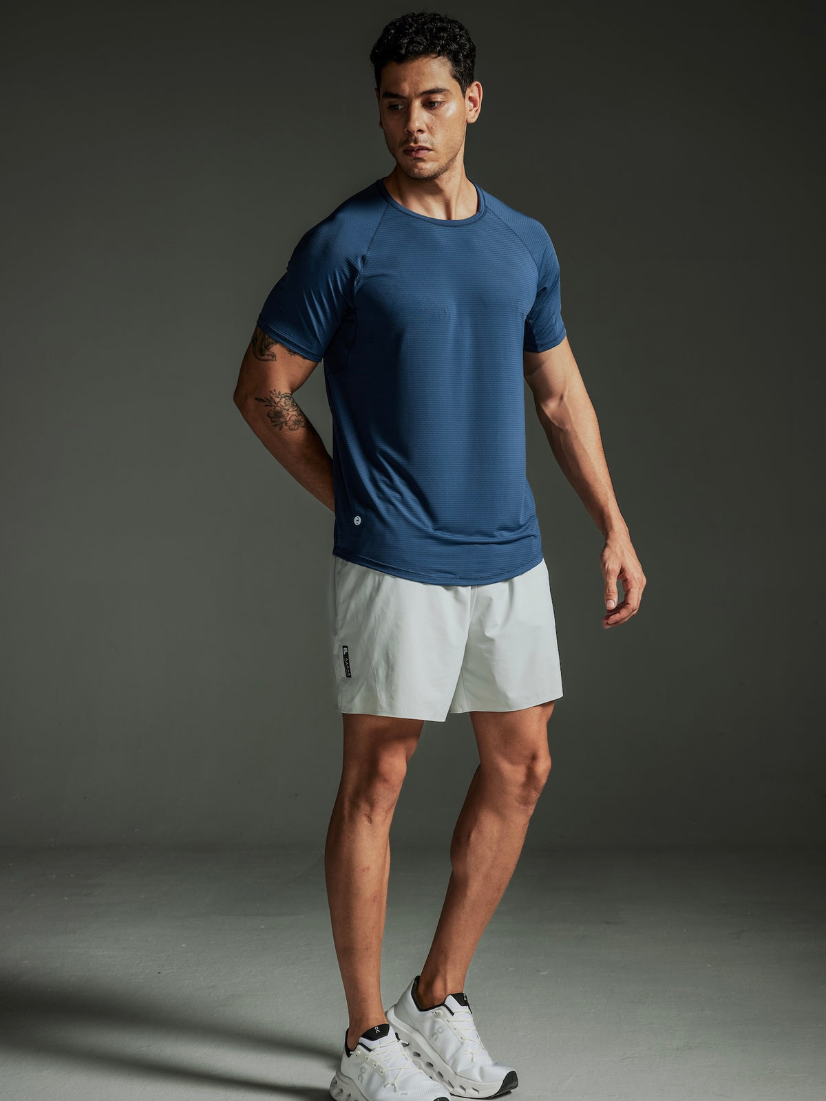 Man wearing a TECHTEE blue athletic shirt and light gray shorts on a dark background