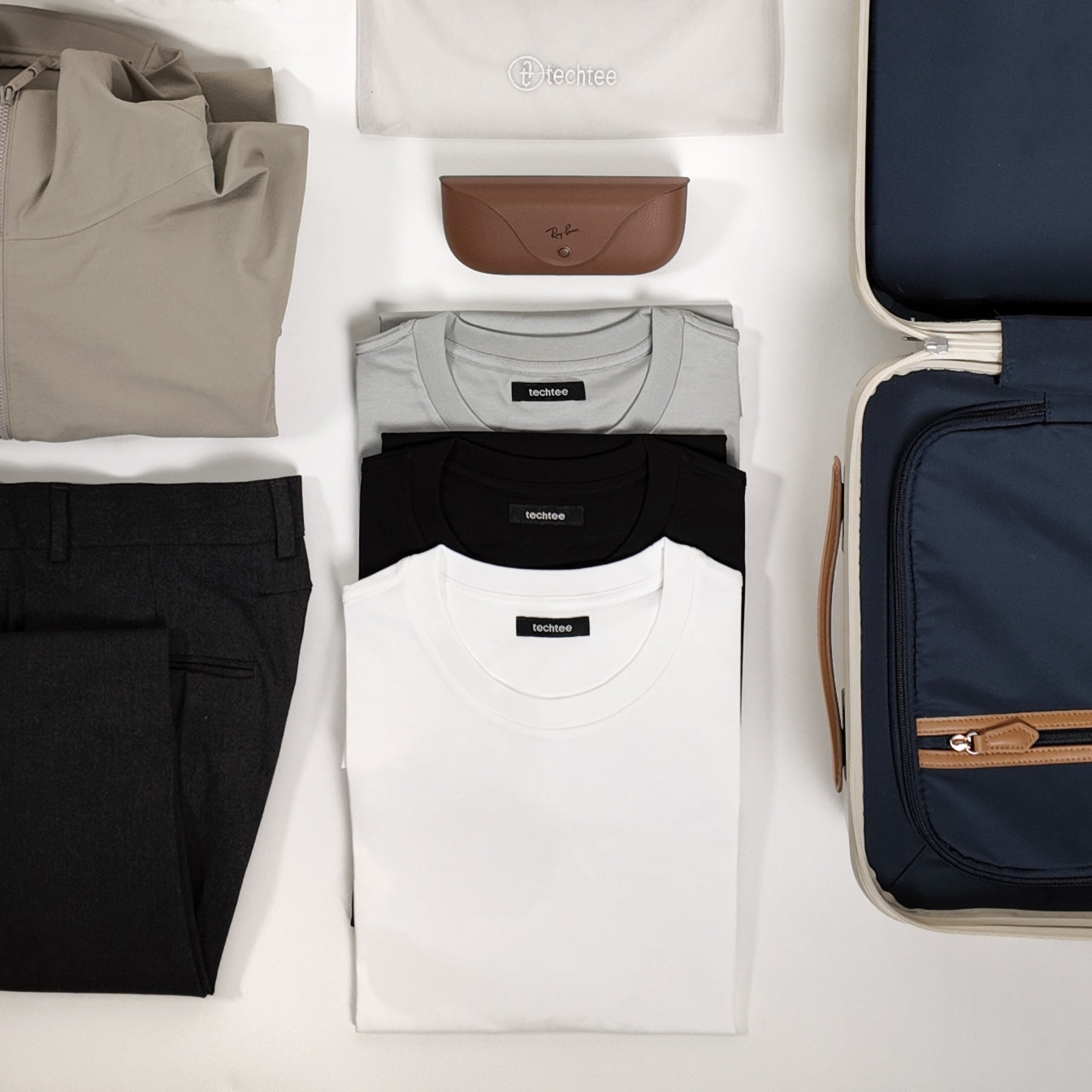 Flat lay of TechTee travel essentials — white, black, and grey classic fit T-shirts neatly folded alongside travel trousers, a packing cube, and carry-on luggage